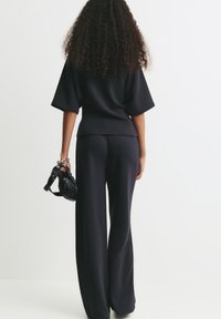 Femme avec de longs cheveux bouclés portant un pantalon large noir et un haut noir ample, tenant un petit sac à main noir à ses côtés.