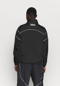 Chaqueta negra con ribetes blancos, cuello alto y etiqueta de logo en la parte posterior. Confeccionada en tela suave; cuenta con puños y cinturilla elásticos.