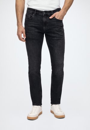 Homme portant un jean noir ajusté, une main dans la poche, et des baskets blanches avec semelles en gomme sur fond blanc.