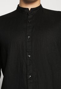 Camicia nera con bottoni, realizzata in un tessuto strutturato, caratterizzata da un colletto alla mandarino e sei bottoni lucidi lungo la patta anteriore.