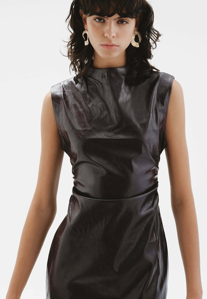 Vestido sin mangas de cuero sintético negro con cuello alto, silueta ajustada y un brillo sutil. Cuenta con una textura suave y detalle de fruncido lateral.