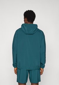 Teal lättviktsjacka med huva, elastiska muddar och nederkant. Bärs över matchande shorts och visar en slät, matt yta.