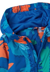 Veste de pluie légère, bleue avec des accents orange ; arbore un motif de dinosaure, capuche élastique et fermeture éclair à l'avant. L'intérieur est doublé en maille.