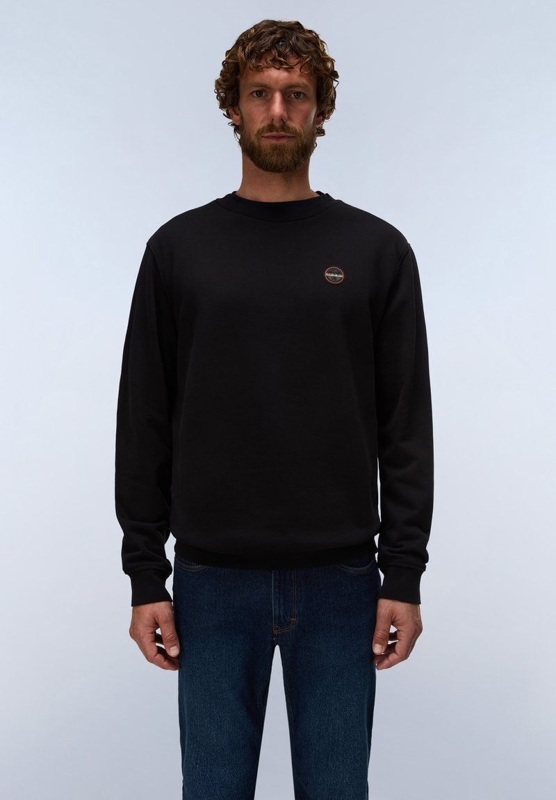 Mand med krøllet hår og skæg iført en sort langærmet sweatshirt og blå jeans, stående mod en ensfarvet lys baggrund.