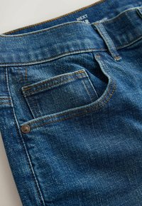 Gros plan sur un jean bleu en denim montrant la poche avant, la poche pour monnaie, les passants de ceinture et les coutures orange visibles sur le tissu texturé.