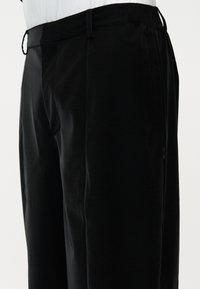 Μαύρο παντελόνι με ίσια γραμμή, ελαστική μέση και θηλιές για ζώνη, φορεμένο με λευκό τοπ, εμφανιζόμενο από τη μέση έως το μέσο του μηρού, πλάγια όψη.