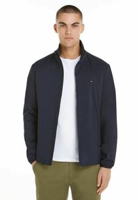 Giacca blu navy con cerniera realizzata in materiale leggero, con colletto alla coreana e dettaglio del logo bianco sul petto sinistro, abbinata a una camicia bianca.