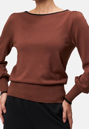 Femme portant un pull marron ajusté avec des manches longues bouffantes et une bordure noire au niveau de l'encolure et des poignets, assorti à un pantalon noir.