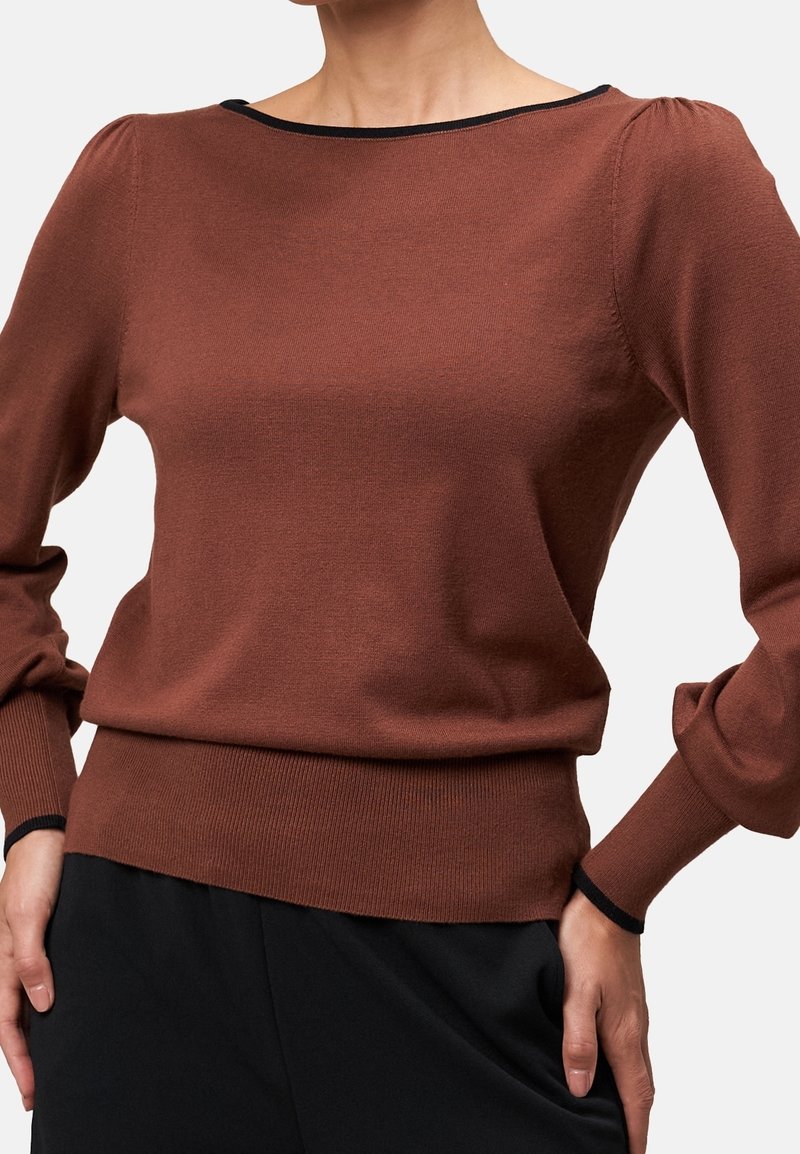 Femme portant un pull marron ajusté avec des manches longues bouffantes et une bordure noire au niveau de l'encolure et des poignets, assorti à un pantalon noir.