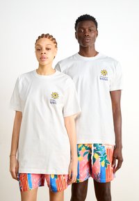 T-shirts de algodão brancas com um design gráfico colorido de um sol e ondas no lado esquerdo do peito, combinadas com calções vibrantes e estampados.