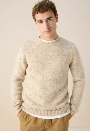Jonge man met krullend haar in een beige spikkelige gebreide trui over een wit shirt en een bruine corduroy broek, handen in de zakken, neutrale achtergrond.