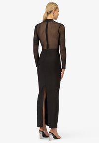 Robe noire à manches longues avec un dos et des manches transparentes, silhouette ajustée, fente sur le côté et fermeture éclair au cou.