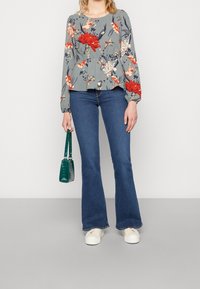 Blusenshirt mit floralem Peplum und langen Ärmeln in Grau, mit roten und weißen Blumen; kombiniert mit blauen Schlagjeans und einer grünen Handtasche.