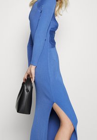 Blå ribbad långärmad klänning med sidospringa, som visar en åtsittande silhuett. Modellen håller en liten svart handväska.