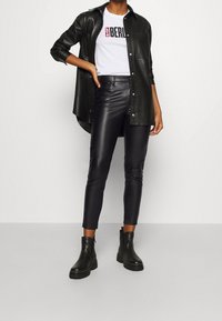 Camicia in pelle nera, t-shirt grafica bianca con la scritta "BERLIN", pantaloni neri attillati e stivali neri alla caviglia con suola spessa. Texture lisce in tutto.