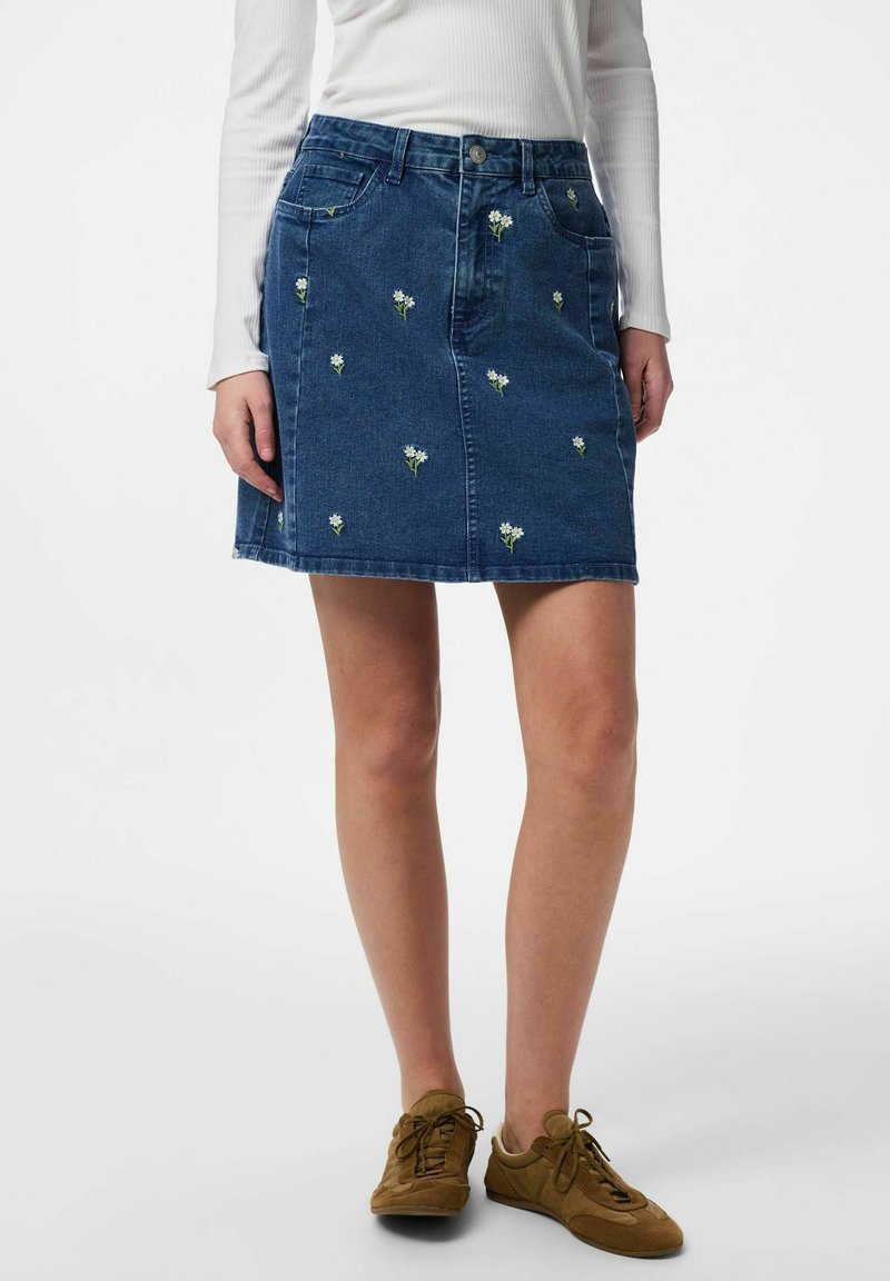 Jupe en denim bleu avec petite broderie de fleurs blanches, portée avec un haut blanc à manches longues et des chaussures marron à lacets, posée devant un fond blanc.