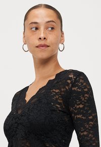 Schwarzes Spitzentop mit V-Ausschnitt und langen Ärmeln, das mit floralen Mustern und einer transparenten Textur versehen ist. Sichtbare hängende Ohringe.