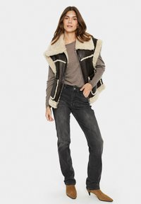 Luxus vest med shearling i mørkebrunt læder, cremet kunstpelsfoer og store revers. Sammen med grå jeans og beige hælede ankelstøvler.