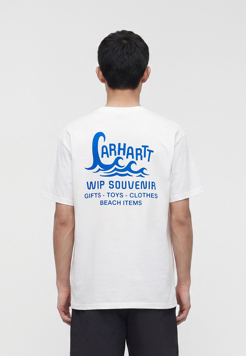 Persona che indossa una t-shirt bianca con la scritta blu "Carhartt WIP Souvenir" e grafiche a onde sulla schiena, abbinata a pantaloncini neri.