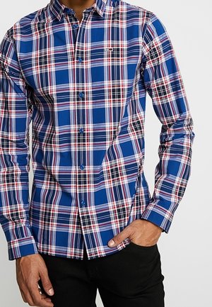 Mann trägt ein blau, rot, weiß und schwarz kariertes Hemd mit langen Ärmeln, kombiniert mit schwarzen Hosen, Hand in der Tasche.