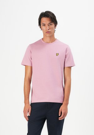 Mann iført en lys rosa t-skjorte med kort erm og gul logo på brystet, og mørke bukser, stående mot en hvit bakgrunn.