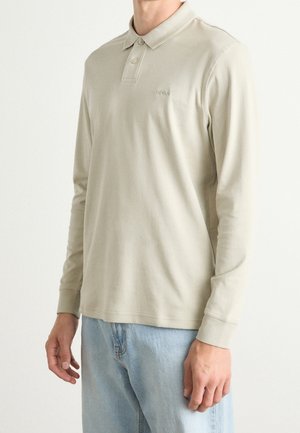 Mężczyzna w beżowej, długorękawowej koszulce polo z zapiętym kołnierzykiem na guziki oraz jasnoniebieskich dżinsach na jednolitym tle.