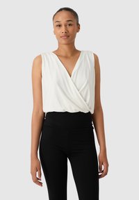 Femme portant un top enroulé sans manches blanc et un legging noir taille haute, se tenant contre un fond gris clair uni.