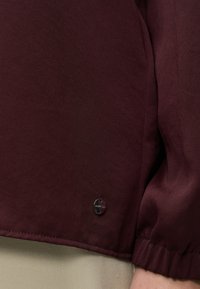 Gros plan d’une personne portant un vêtement à manches longues bordeaux foncé avec un poignet élastique et un petit logo métallique rond près de l’ourlet.