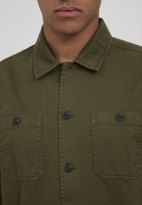 Olive green shirt jacket made of cotton, featuring a collared neck, two chest pockets with black buttons, and a textured fabric finish.