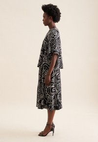 Abito nero con motivo floreale bianco, design svasato, maniche corte e orlo knee-length. Indossato con sandali neri con tacco alto.