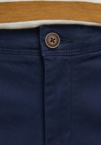 Tummansiniset housut, joissa on sileä pinta, ruskea nappi, ompeluyksityiskohtia ja päällä taitettu beige ja ruskea raidallinen paita.