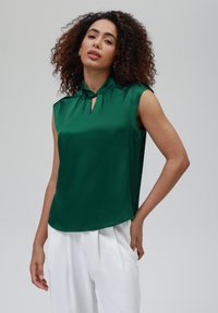 Blusa senza maniche verde smeraldo con scollatura a foro e dettagli arricciati. Tessuto liscio e lucido. Abbinata a pantaloni bianchi plissettati.