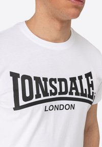 Bílý bavlněný tričko s výrazným černým logem "LONSDALE" s zakřiveným designem a textem "LONDON" pod ním, umístěné na hladké látce.