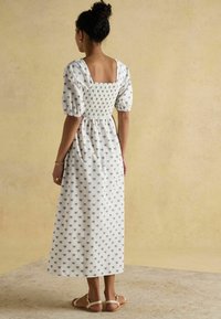 Rochie midi albă cu imprimeu floral albastru, spate pătrat, corset elastic și mâneci bufante. Pur tată cu sandale plate bej. Fundal texturat bej.