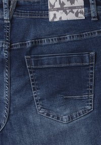 Jean en denim avec une teinte bleu foncé, des coutures visibles et une poche arrière. L'étiquette comprend un motif abstrait en gris et blanc.