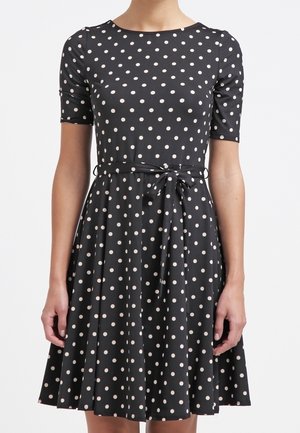 Robe noire longueur genou avec des pois blancs, manches courtes, encolure ronde et ceinture nouée à la taille, portée par une personne debout.