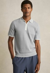 Polo shirt scurt, de culoare gri deschis, cu textură tricotată în cablu, guler cu fermoar și detalii contrastante albe. Asortat cu pantaloni în dungi închise.