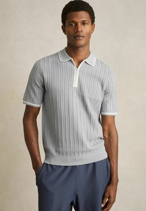 Polo shirt scurt, de culoare gri deschis, cu textură tricotată în cablu, guler cu fermoar și detalii contrastante albe. Asortat cu pantaloni în dungi închise.