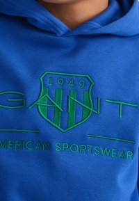 Blå hoodie i bomull med en broderad grön sköldlogotyp med året "1949" och texten "GANT AMERICAN SPORTSWEAR."