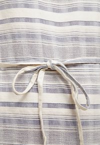 Kevyt puuvillainen esiliina, jossa on vaaleansiniset ja valkoiset vaakaraitat, varustettuna solmittavalla vyötäröllä. Rikkaita tekstuureja tuovat visuaalista kiinnostavuutta.