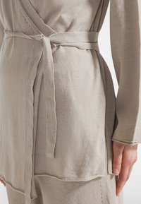 Béžový zavinovací top s texturovanou látkou, páskem na zavázání v pase a prošitými detaily na lemu. Má dlouhé rukávy a boční rozparek.