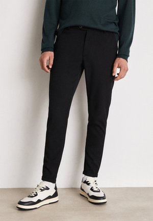 Hombre vestido con pantalones ajustados negros, zapatillas blancas y negras, y una camiseta de manga larga verde oscuro, de pie contra una pared lisa y clara.