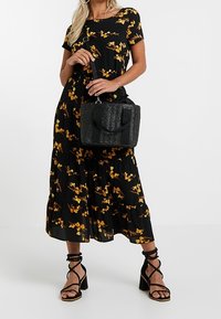 Vestido largo negro con flores amarillas, combinado con un bolso negro texturizado y sandalias negras con tiras y un tacón ancho de madera.