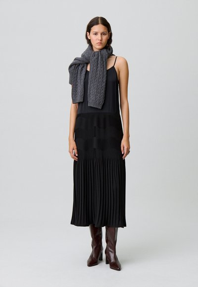 Vestito nero plissettato che arriva a metà polpaccio, abbinato a una sciarpa grigia lavorata a maglia. Il modello indossa stivali marroni con tacco. Tessuto liscio con contrasto di texture sottile.