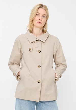 Blonde Frau trägt beige, geknöpfte Jacke mit großem Kragen und hellblaue Jeans, steht mit den Händen in den Taschen vor weißem Hintergrund.