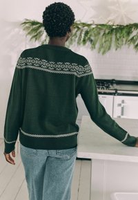 Dunkelgrüner Pullover mit einem dekorativen weißen Muster über den Schultern und am Saum; kombiniert mit hellblauen Jeans in einer Küchenszenerie.