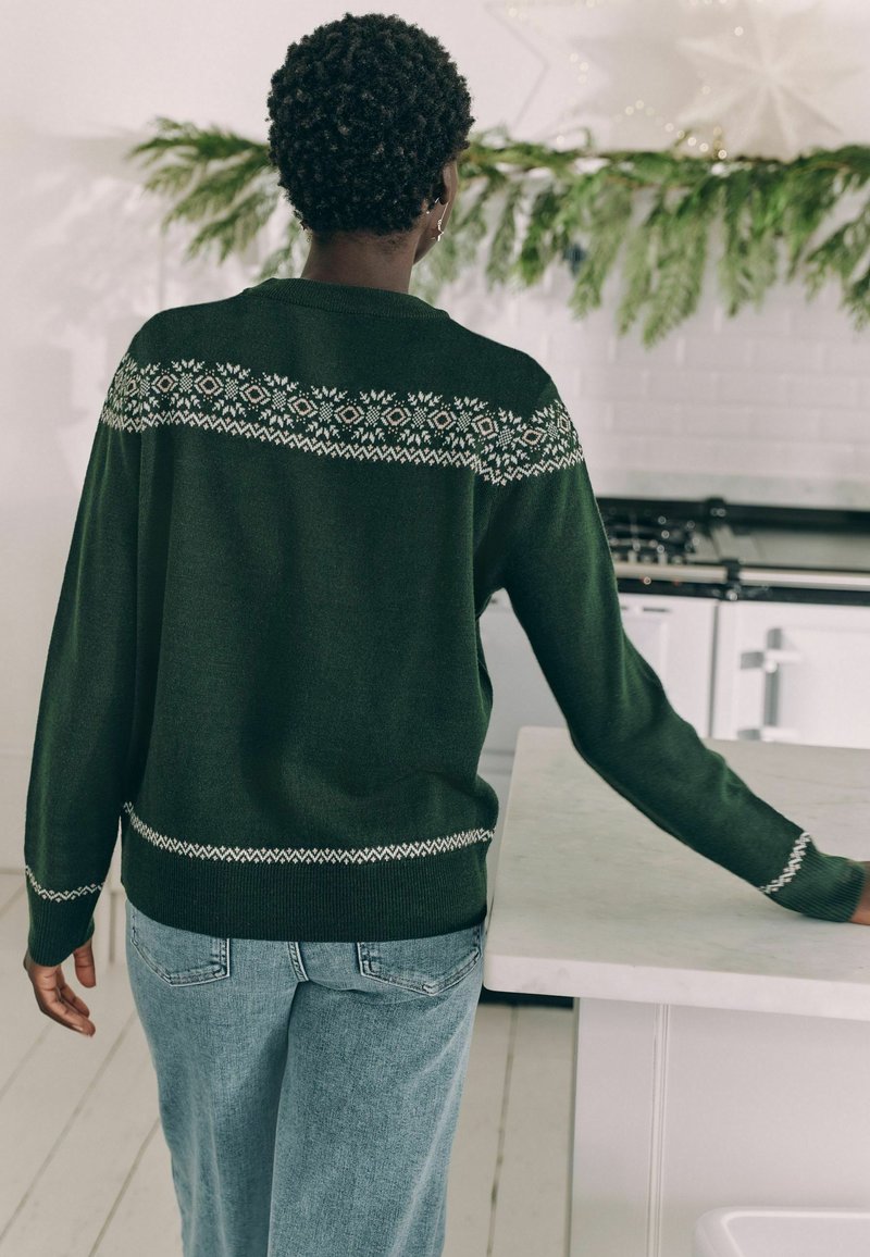 Dunkelgrüner Pullover mit einem dekorativen weißen Muster über den Schultern und am Saum; kombiniert mit hellblauen Jeans in einer Küchenszenerie.