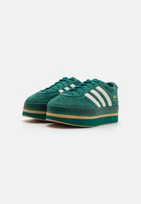 Sapatilhas de plataforma em suede verde com três riscas brancas, bico redondo e sola de borracha texturizada com detalhe em risca bege. Acento em dourado na marca.