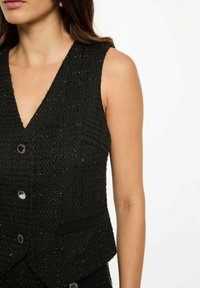 Donna che indossa un gilet nero senza maniche con bottoni argentati e pantaloni abbinati su uno sfondo bianco.