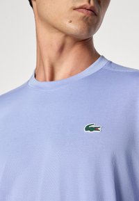 Ανοιχτό μπλε t-shirt με στρογγυλή λαιμόκοψη, φτιαγμένο από μαλακό βαμβάκι. Διαθέτει ένα μικρό πράσινο λογότυπο κροκόδειλου στο αριστερό στήθος. Ομαλή υφή, άνετη εφαρμογή.